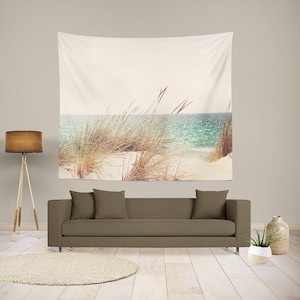 May include: A tapestry featuring a beach scene with tall grass in the foreground and a blue ocean in the background. The sky is a light blue and the sand is white.