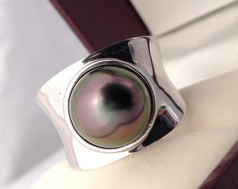 Könnte beinhalten: Ein silberner Ring mit einer großen, schillernden Perle, die in einem modernen, geometrischen Design gefasst ist. Die Perle zeigt eine Reihe von Farben, darunter Grün, Lila und Schwarz. Der Ring wird in einer burgunderfarbenen Schmuckschatulle mit cremefarbenem Innenraum präsentiert.