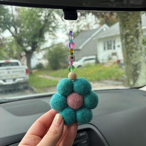Peut inclure: Une décoration de voiture suspendue composée d'une fleur en feutrine avec des pétales bleu sarcelle et un centre rose. La fleur est fixée à un cordon de perles colorées, incluant un petit œil porte-bonheur, une perle violette et une verte.