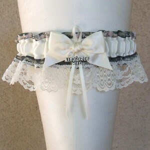 May include: A white garter with a cream-colored bow and the text "AIR FORCE WIFE". The garter features a lace trim and a patterned band with gray and purple accents. The bow has long, cream-colored ribbons.