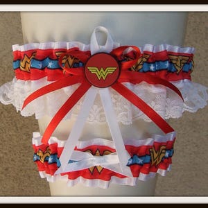 May include: A set of two leg garters featuring a red, blue, and white ribbon pattern with gold Wonder Woman logos and white stars. The garters are trimmed with white lace and decorated with red and white satin ribbons and a central circular emblem.