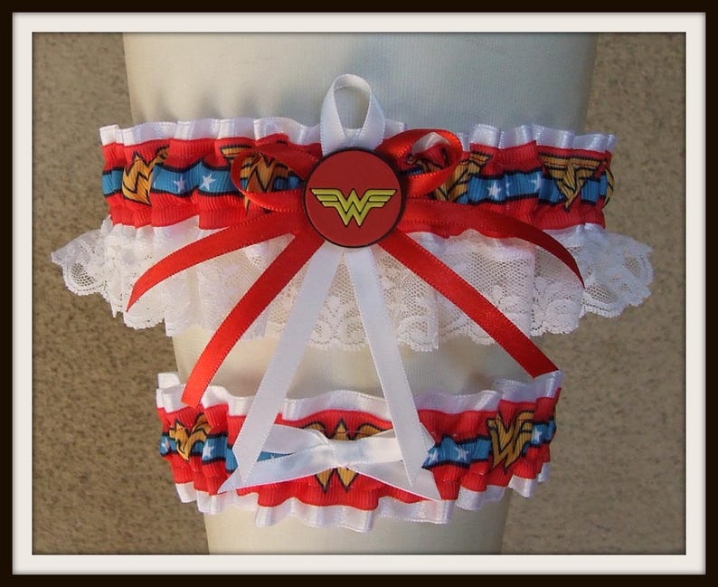 May include: A set of two leg garters featuring a red, blue, and white ribbon pattern with gold Wonder Woman logos and white stars. The garters are trimmed with white lace and decorated with red and white satin ribbons and a central circular emblem.