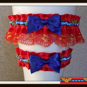 May include: A set of two red elastic garters featuring a blue ribbon with white stars and gold lightning bolt patterns. Each garter is adorned with a deep blue satin bow and a layer of red lace with gold metallic thread accents throughout.