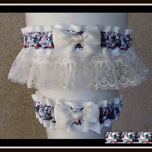 May include: A set of two white garters featuring a patterned fabric with red and teal accents, topped with a solid white satin bow. The larger garter includes a wide layer of intricate white lace trim, while both pieces have a gathered elastic band.