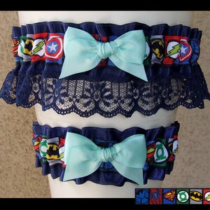 May include: Two elastic leg garters featuring a colorful superhero comic book print with various character emblems. Each garter is accented with a solid light blue satin bow and dark blue ruffled trim, with the top garter featuring additional black lace.