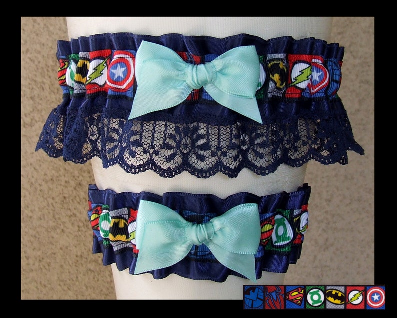 May include: Two elastic leg garters featuring a colorful superhero comic book print with various character emblems. Each garter is accented with a solid light blue satin bow and dark blue ruffled trim, with the top garter featuring additional black lace.
