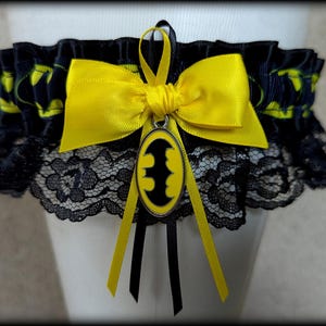 May include: A black garter featuring a yellow and black Batman logo pattern on the ribbon, accented with a bright yellow satin bow and a central oval charm displaying a black bat symbol on a yellow background, finished with black lace trim.
