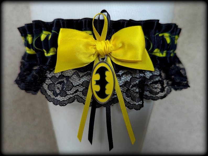 May include: A black garter featuring a yellow and black Batman logo pattern on the ribbon, accented with a bright yellow satin bow and a central oval charm displaying a black bat symbol on a yellow background, finished with black lace trim.
