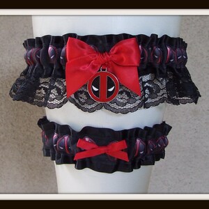 May include: A set of two black elastic garters featuring a repeating Deadpool mask pattern. The larger garter includes a red satin bow and a circular Deadpool logo charm, while the smaller garter features a simple red satin bow accent.