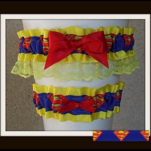 May include: A set of two elastic garters featuring a blue ribbon with a red and yellow Superman logo print. The garters are trimmed with yellow ruffles and a yellow lace border, each accented with a centered red satin bow on the front.