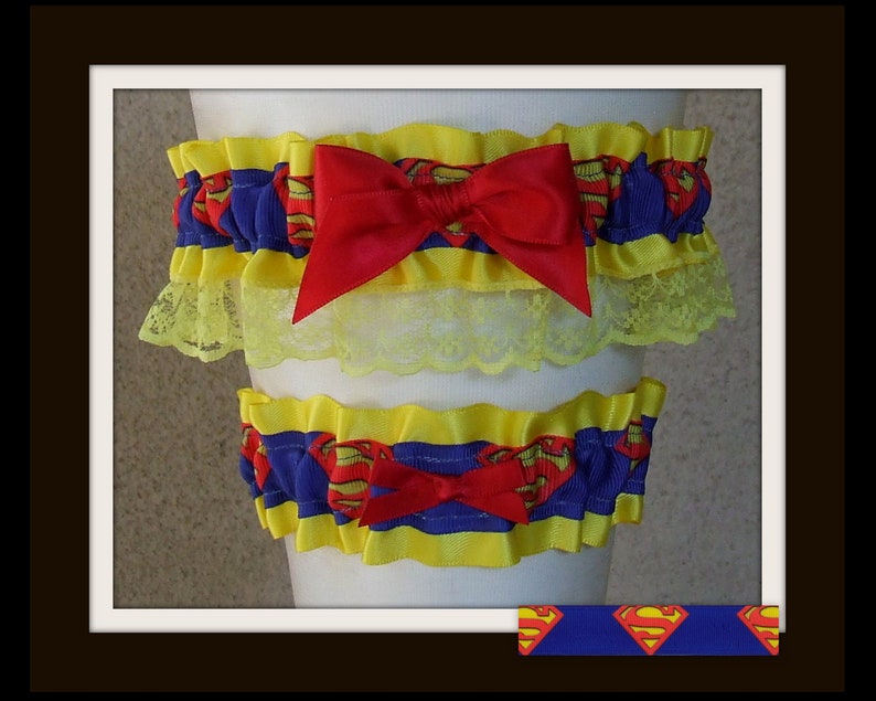 May include: A set of two elastic garters featuring a blue ribbon with a red and yellow Superman logo print. The garters are trimmed with yellow ruffles and a yellow lace border, each accented with a centered red satin bow on the front.