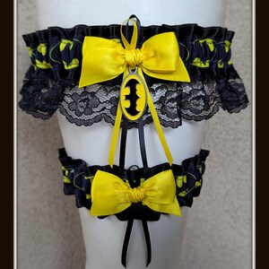 May include: A set of two black and yellow garters featuring a repeating bat logo pattern. The top garter includes black lace trim, a yellow satin bow, and a central oval charm with a black bat silhouette. A matching smaller garter has a yellow bow.