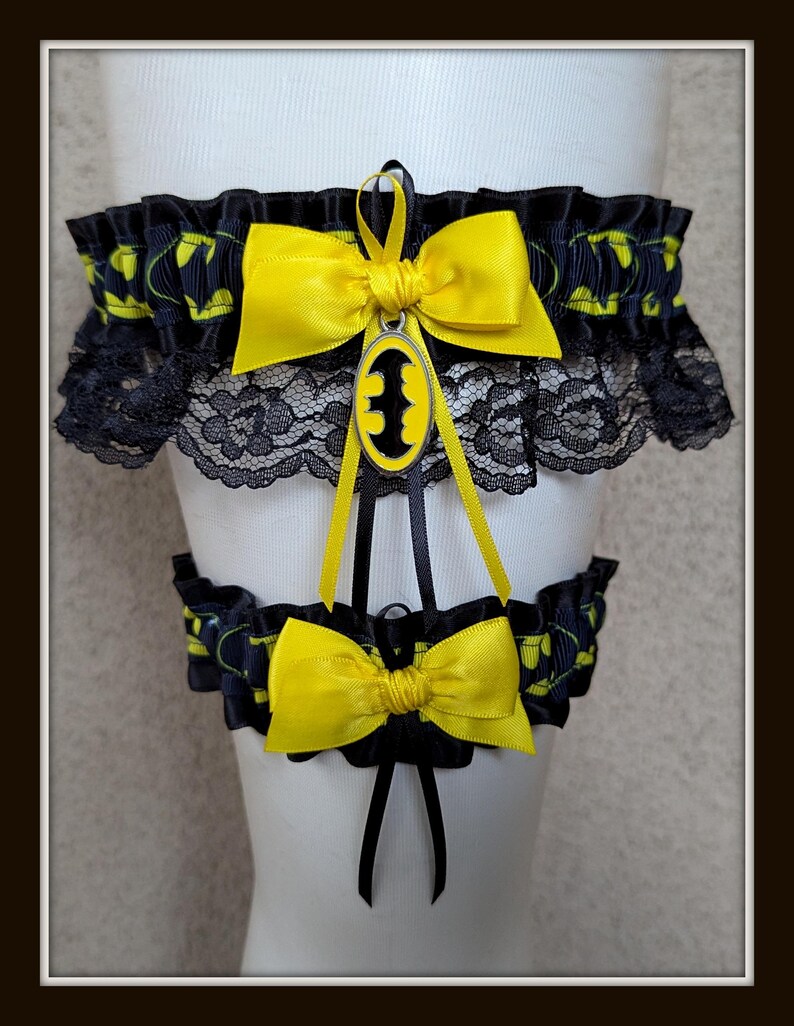 May include: A set of two black and yellow garters featuring a repeating bat logo pattern. The top garter includes black lace trim, a yellow satin bow, and a central oval charm with a black bat silhouette. A matching smaller garter has a yellow bow.