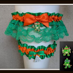 May include: A set of two elastic garters featuring green lace and orange satin ribbon trim. The larger garter has an orange satin bow with a metal charm of a green turtle character. The smaller garter features a silver-colored pizza slice charm.