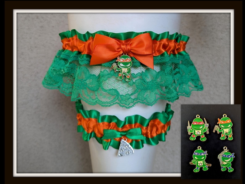 May include: A set of two elastic garters featuring green lace and orange satin ribbon trim. The larger garter has an orange satin bow with a metal charm of a green turtle character. The smaller garter features a silver-colored pizza slice charm.