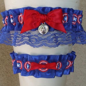 May include: A set of two blue elastic garters featuring a repeating red, white, and blue shield pattern. The larger garter includes a red satin bow with a silver-toned star charm, while the smaller garter features a simple red satin bow.