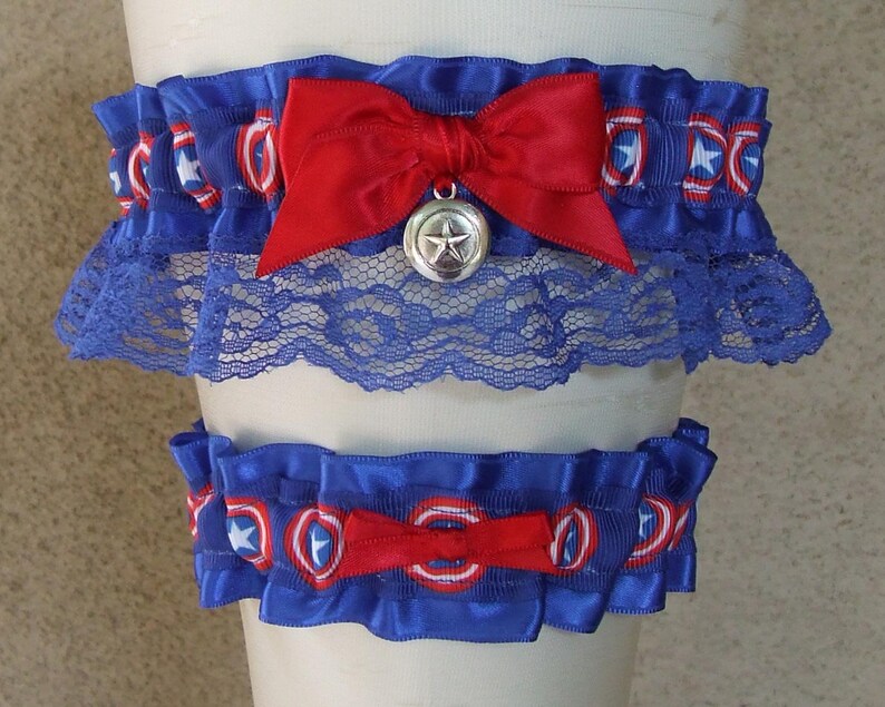 May include: A set of two blue elastic garters featuring a repeating red, white, and blue shield pattern. The larger garter includes a red satin bow with a silver-toned star charm, while the smaller garter features a simple red satin bow.