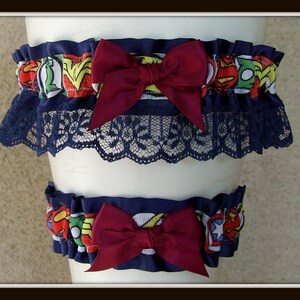 May include: A set of two leg garters featuring a colorful comic book superhero print with red, yellow, blue, and green accents. Each garter is adorned with a deep burgundy satin bow and finished with a dark navy blue lace trim along the bottom edge.