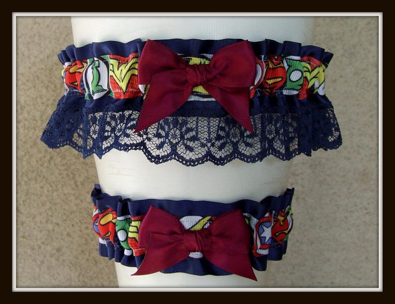 May include: A set of two leg garters featuring a colorful comic book superhero print with red, yellow, blue, and green accents. Each garter is adorned with a deep burgundy satin bow and finished with a dark navy blue lace trim along the bottom edge.
