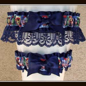 May include: A set of two leg garters featuring a navy blue satin ribbon bow and a navy blue lace trim. The elasticized bands are decorated with a colorful comic book character print pattern, showcasing various superhero faces in a repeating design.