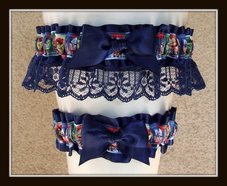 May include: A set of two leg garters featuring a navy blue satin ribbon bow and a navy blue lace trim. The elasticized bands are decorated with a colorful comic book character print pattern, showcasing various superhero faces in a repeating design.
