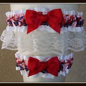 May include: A set of two leg garters featuring a white fabric base with a red and blue spiderweb pattern. Each garter is adorned with a large, bright red satin bow and a wide layer of white floral lace trim along the bottom edge of the top piece.