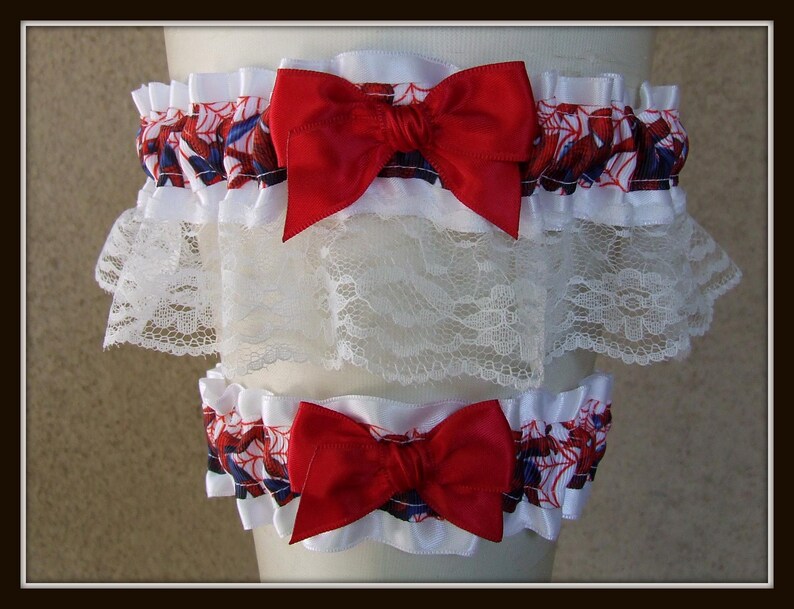 May include: A set of two leg garters featuring a white fabric base with a red and blue spiderweb pattern. Each garter is adorned with a large, bright red satin bow and a wide layer of white floral lace trim along the bottom edge of the top piece.