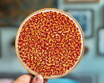 May include: A close-up of a round embroidery hoop with a red and orange floral design. The hoop is being held by a hand.