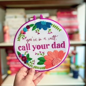 May include: Embroidered hoop with a white background and colorful flowers. The text "you're in a cult, call your dad" is embroidered in pink.