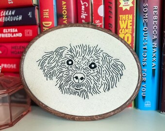 May include: A black and white embroidered image of a dog's face on a white canvas in a wooden embroidery hoop.