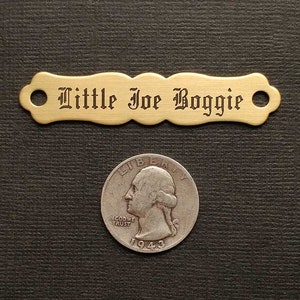 Może przedstawiać: Metalowa blaszka z grawerem "Little Joe Boggie" w ozdobnym stylu pisma. Blaszka ma po dwa otwory na każdym końcu.
