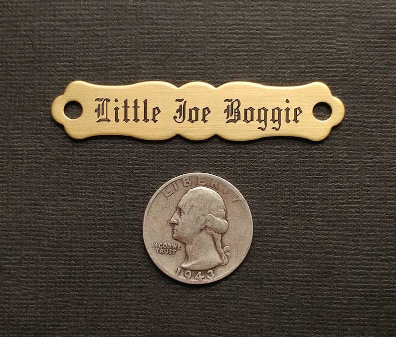 Może przedstawiać: Metalowa blaszka z grawerem "Little Joe Boggie" w ozdobnym stylu pisma. Blaszka ma po dwa otwory na każdym końcu.