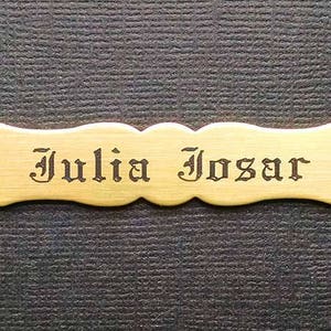 Może przedstawiać: Mosiężna tabliczka z falistym brzegiem i dwoma otworami do zawieszenia. Imię "Julia Josar" jest wygrawerowane czarną czcionką.