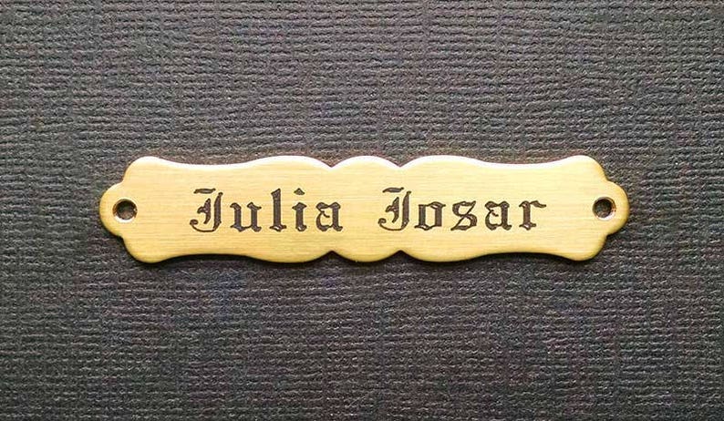 Może przedstawiać: Mosiężna tabliczka z falistym brzegiem i dwoma otworami do zawieszenia. Imię "Julia Josar" jest wygrawerowane czarną czcionką.