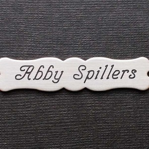 Może przedstawiać: Srebrna tabliczka z imieniem o zakrzywionym kształcie i zaokrąglonych krawędziach. Imię "Abby Spillers" jest wygrawerowane ozdobnym pismem.