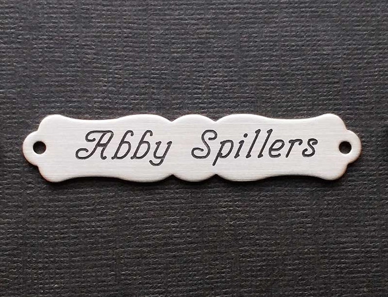Może przedstawiać: Srebrna tabliczka z imieniem o zakrzywionym kształcie i zaokrąglonych krawędziach. Imię "Abby Spillers" jest wygrawerowane ozdobnym pismem.