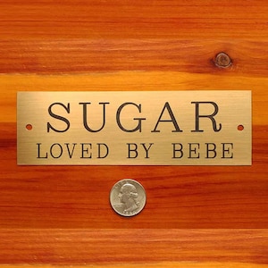 Puede incluir: Un letrero de metal dorado con el texto "SUGAR. LOVED BY BEBE." sobre una superficie de madera.