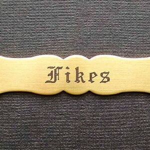 Może przedstawiać: Mosiężna tabliczka z wygrawerowanym napisem "Fikes" w stylu gotyckim.
