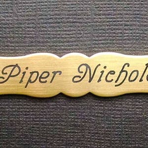 Może przedstawiać: Metalowa tabliczka z nazwiskiem w kolorze złotym z wygrawerowanym czarnym tekstem "Piper Nichole".