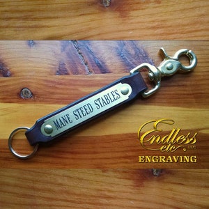 May include: A brown leather keychain with a brass tag engraved with the text "Mane Steed Stables". The keychain has a brass key ring and a brass swivel clasp.