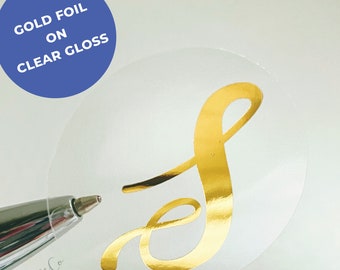 May include: A gold foil letter S on a clear gloss sticker. The sticker is round and has a white background.