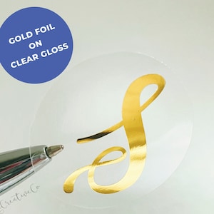 May include: A gold foil letter S on a clear gloss sticker. The sticker is round and has a white background.
