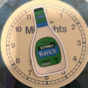 May include: A white and green sticker of a ranch dressing bottle with the text "Seemingly Ranch" on it. The sticker is placed on a clock face with the numbers 1 through 12.