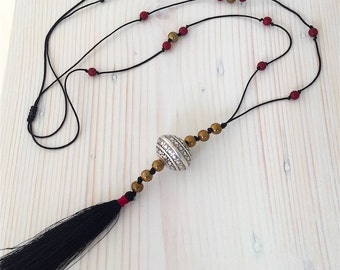 Puede incluir: Un collar largo negro con una cuenta plateada, cuentas doradas y cuentas rojas. El collar tiene una borla negra en el extremo. El collar está sobre una superficie de madera clara.