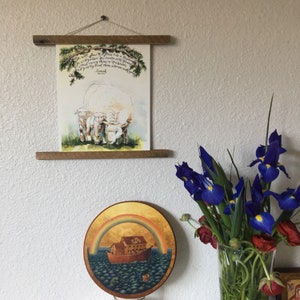 May include: A watercolor painting of three white sheep with a quote from the Bible, "He will feed his flock like a shepherd: He will gather the lambs in his arms and carry them close to his heart; gently will he lead those that are with young."  - Isaiah 40:11. The painting is framed in a wooden frame and hanging on a white wall.