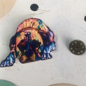 May include: A colorful, abstract, tie-dye style image of a dog's head. The image is printed on a round, white button.