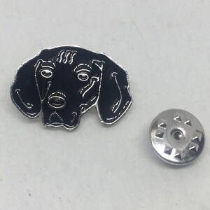 May include: Black and white enamel pin of a dog's head with a silver metal pin back.
