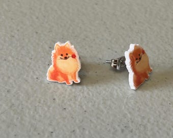 May include: A pair of orange and white stud earrings featuring a cartoon Pomeranian dog with a red flower.