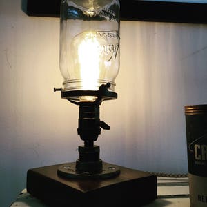 May include: A rustic table lamp made from a repurposed glass jar with a light bulb inside. The lamp has a metal base and a wooden platform.