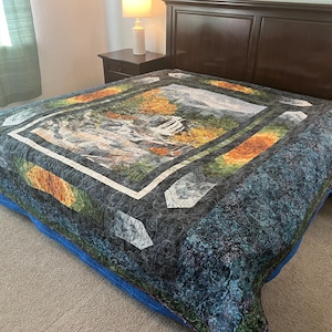 May include: A quilted bedspread featuring a landscape scene with mountains, trees, and water. The quilt has a dark blue background with a repeating pattern, and a blue border. The central image is framed with white and green borders.